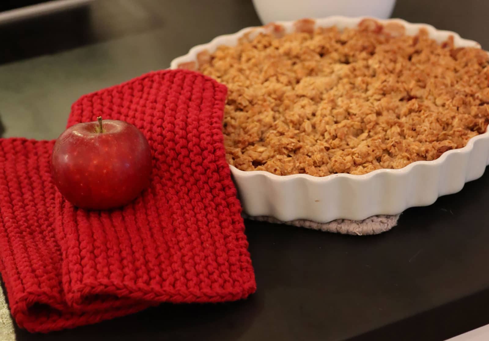 Die Solwang Topflappen in tiefrot liegen neben einem frisch gebackenen Apfelkuchen. Auf den Topflappen ruht ein Apfel.