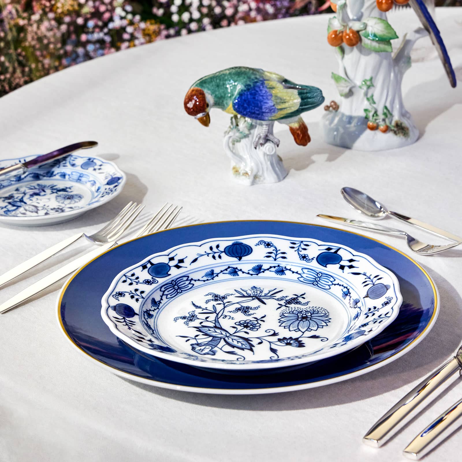 Auf einem gedeckten Tisch steht ein Teller mit dem traditionellen Zwiebelmuster auf dem blauen Urban Colors Speiseteller. Neugierig beugt sich ein bunter Vogel darüber, um ihn zu betrachten.