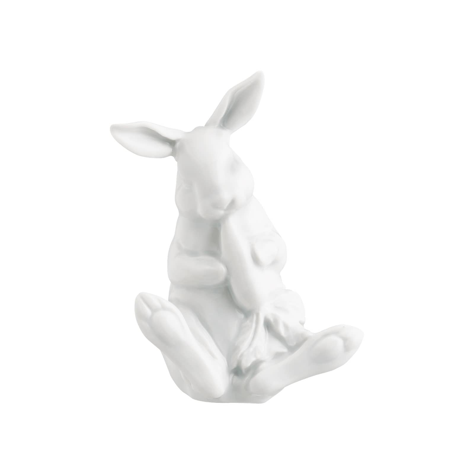 Ein weißer Porzellanhase mit aufgestellten Ohren, der mit beiden Vorderpfoten eine Karotte hält und an ihr knabbert.