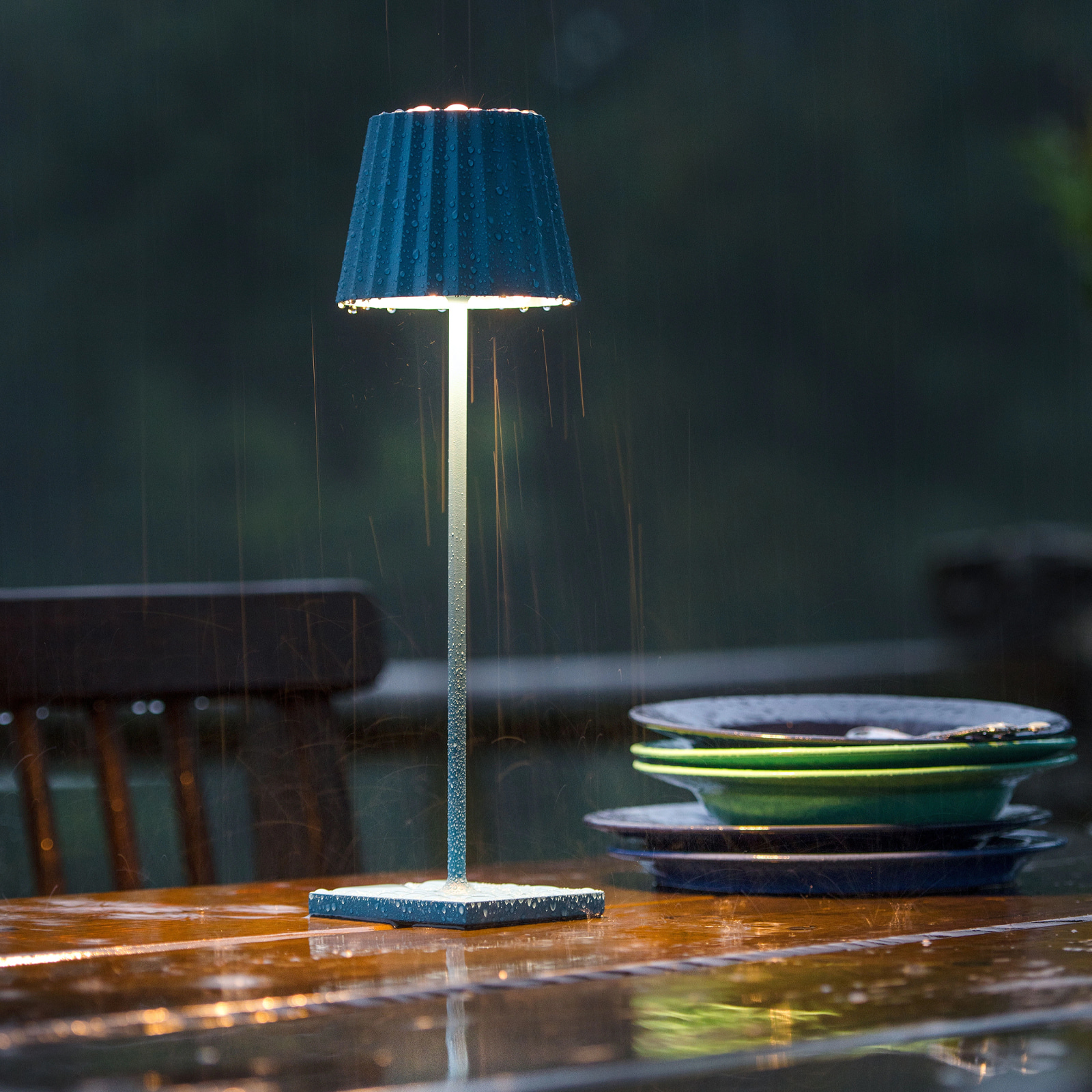 Eine blaue Akku-Tischleuchte auf einem dekorierten Outdoortisch, die im regen steht.