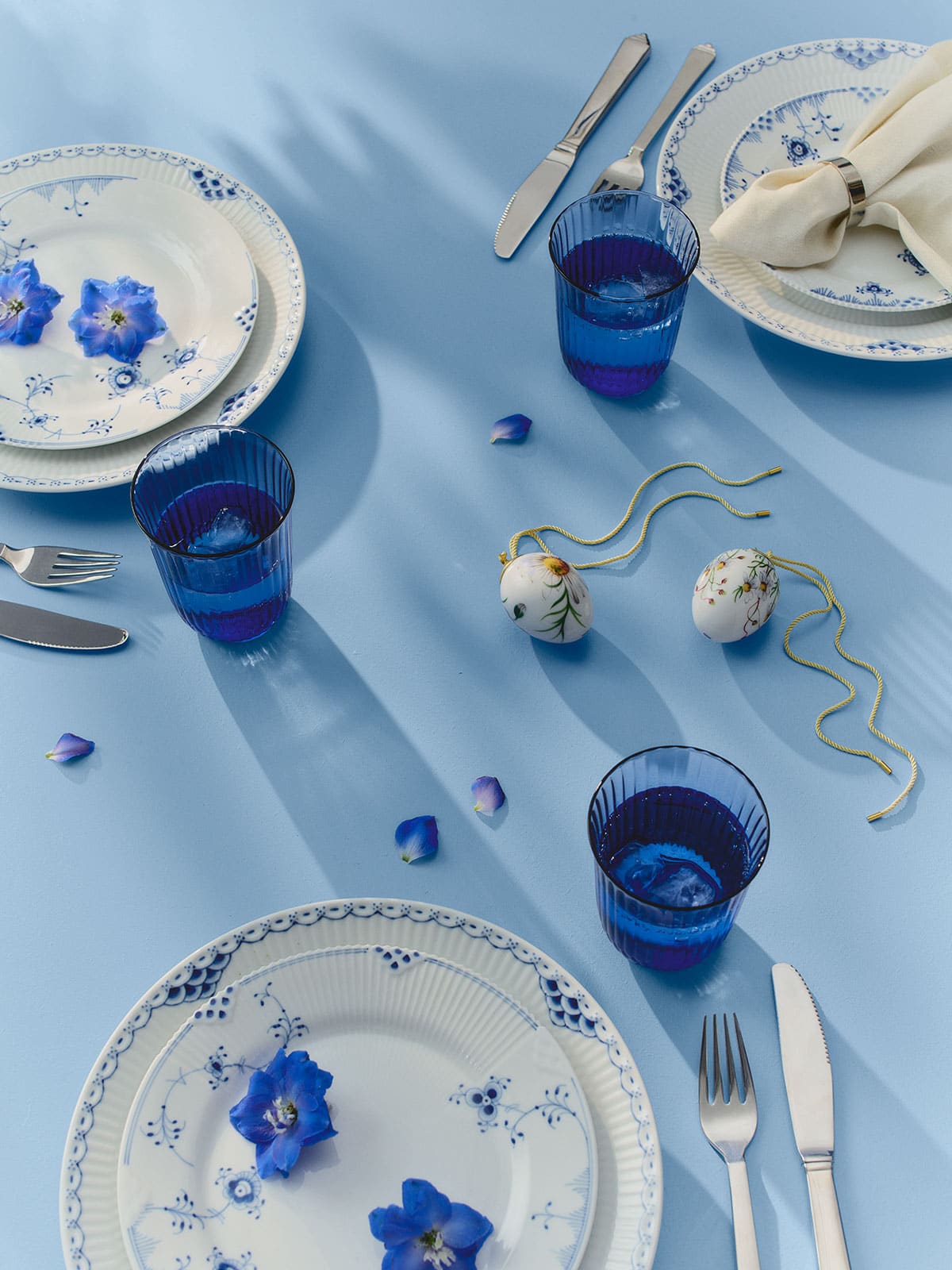 Auf einer hellblauen Tischdecke, zwischen blau dekoriertem Geschirr und kobaltblauen Wassergläsern, liegt das Osterei Kleine Kamille 2026 von Royal Copenhagen.