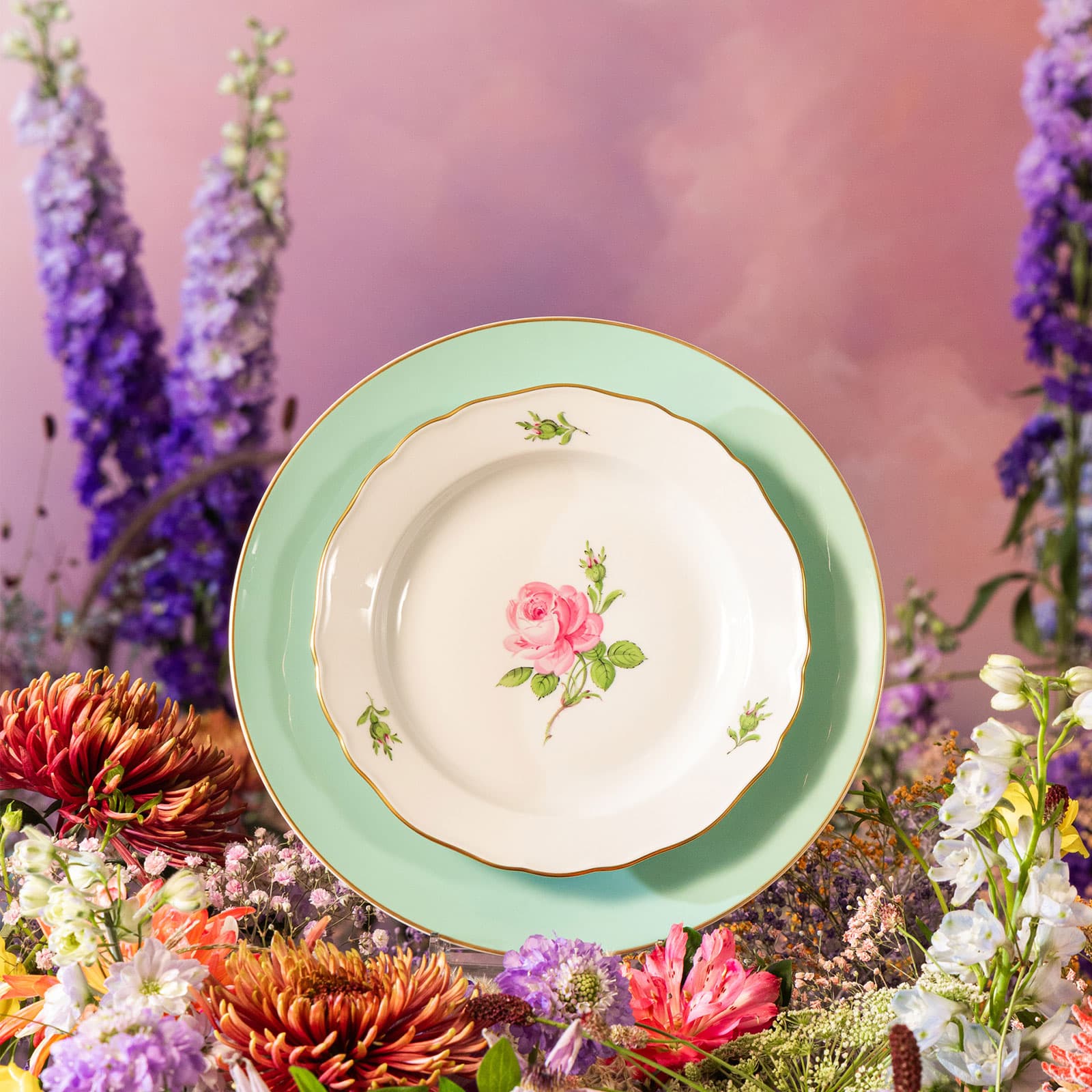 Vor einem Hintergrund aus arrangierten bunten Blumen steht ein Teller des Dekors Meissener Rose auf einem seladongrünen Urban Colors Speiseteller von Meissen.