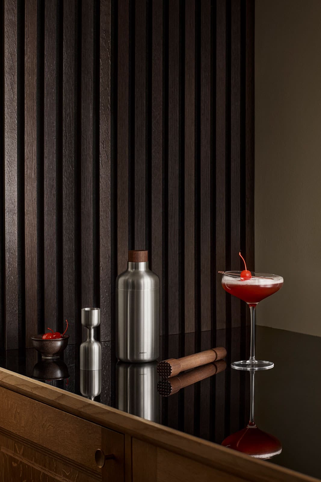 Ein Sideboard bei schwummerigen Licht. Auf der Ablage befinden sich der Cocktailshaker und weitere Artikel der Serie sowie ein gefülltes Cocktailglas.